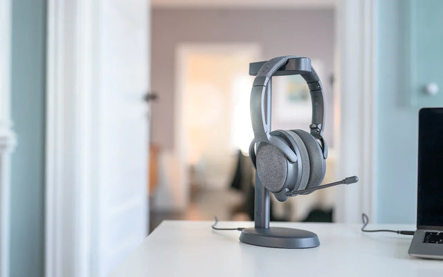 A gray Onanoff Fokus+ ANC headphones with a microphone rests on a sleek stand on a white desk, with a soft-focus background of a stylish room.