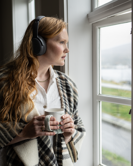 A woman drinking coffee & enjoying music with ONANOFF Fokus+ Active Noise Cancellation Headphones in dusk gray. 
